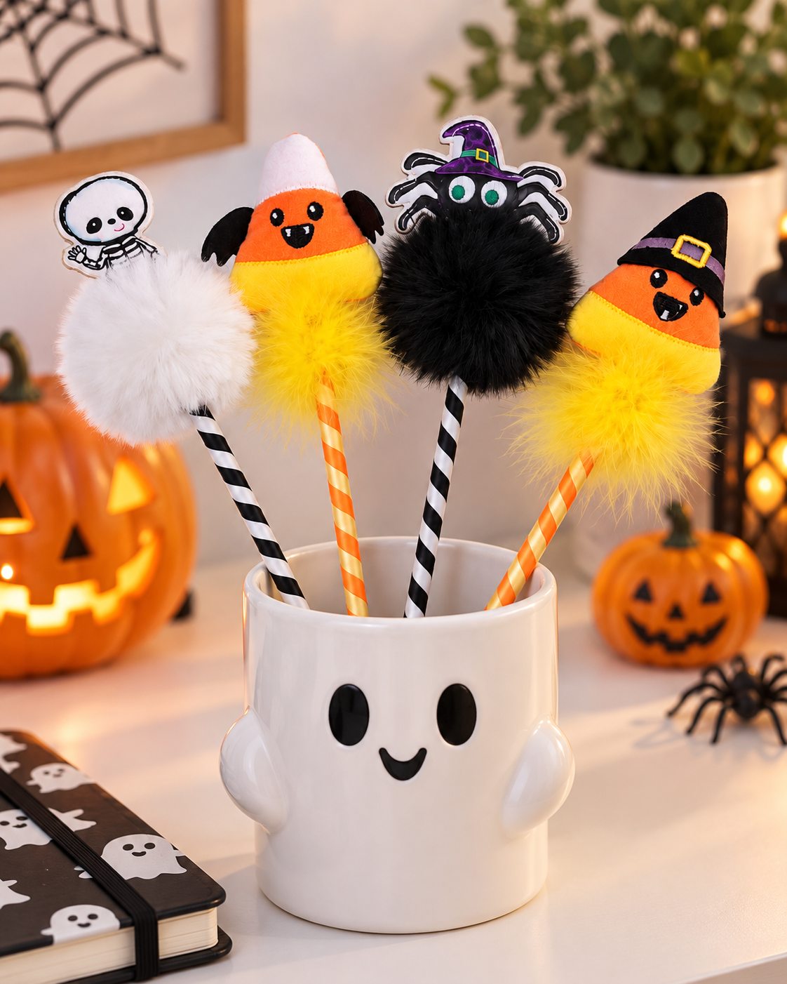 Four Halloween-themed novelty pens with fluffy pom-pom tops are displayed in a white ghost-shaped mug. The pens feature a white skeleton, a yellow-and-orange candy corn, a black spider, and a yellow-and-orange candy corn with a black witch hat.
