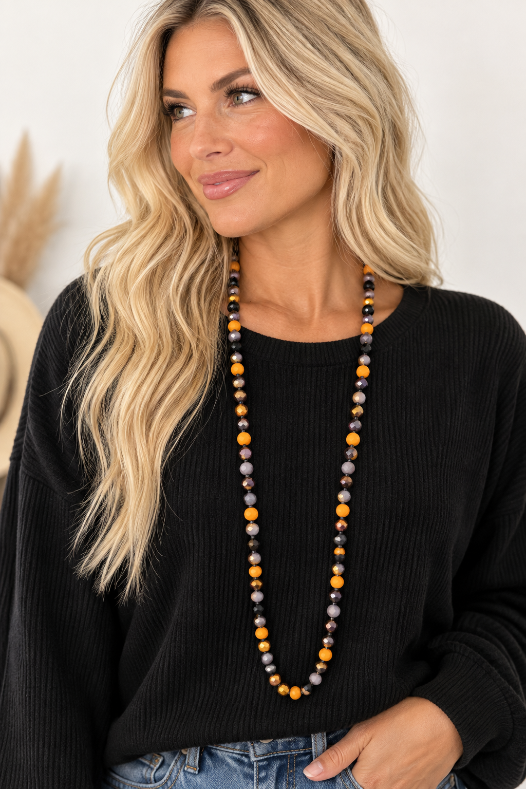 A woman with blonde hair wearing a long beaded necklace with orange, black, and gray beads, paired with a black ribbed sweater and blue jeans.