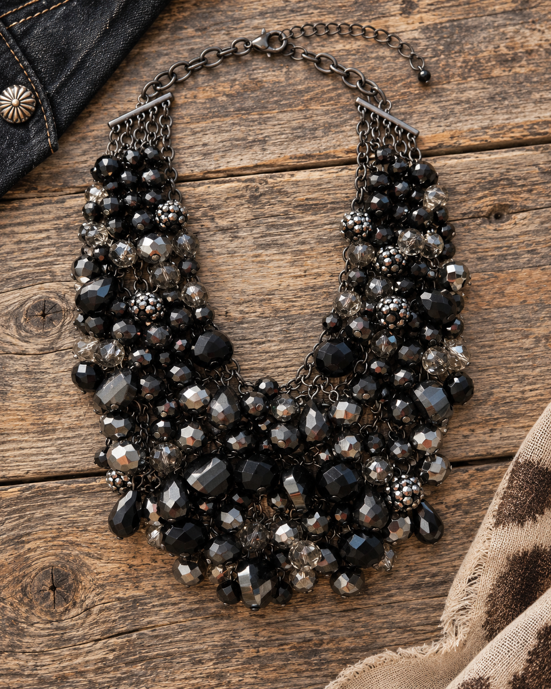A multi-strand statement necklace featuring black, dark gray, and clear faceted beads with an antiqued bronze-tone chain, displayed on a rustic wooden surface.