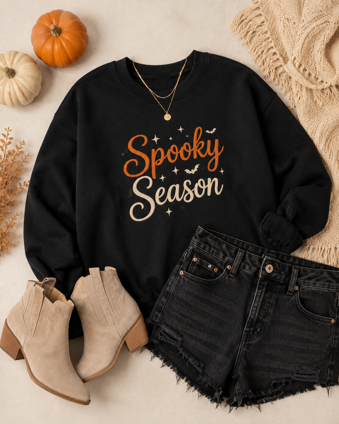 Flat lay of the black Spooky Season embroidered crewneck styled with black denim shorts, neutral ankle boots, gold jewelry, and pumpkins.
