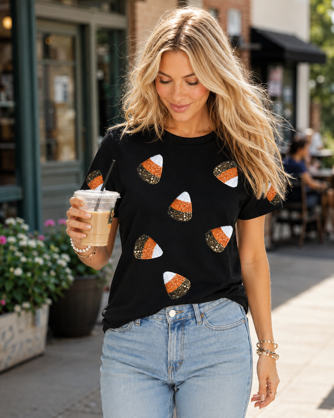 Blonde model wearing the black candy corn sequin Halloween tee with jeans while carrying an iced coffee outside a café.