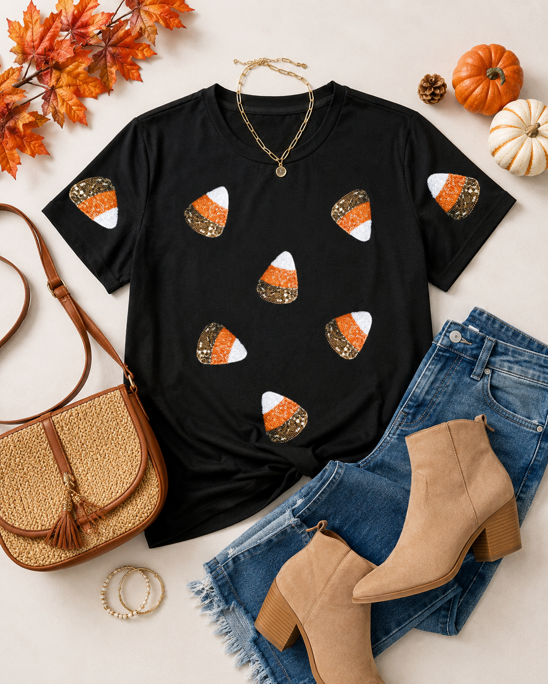 Flat lay of the black sequin candy corn tee with blue denim, tan ankle boots, woven crossbody bag, gold jewelry, pumpkins, and fall leaves.