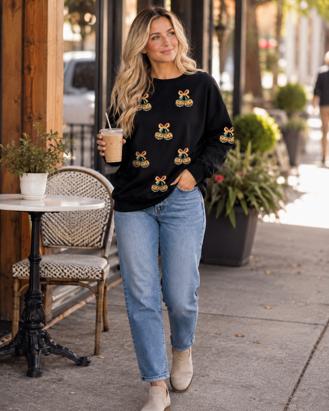 Blonde model styling the black Pumpkin Bow Sequin Sweatshirt with light-wash jeans and iced coffee outside a café.