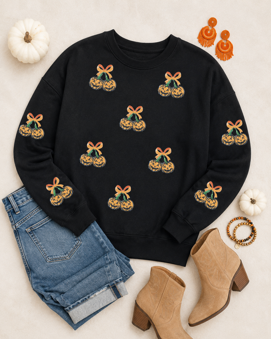Flat lay of the black Pumpkin Bow Sequin Sweatshirt with denim, tan western boots, orange earrings, bracelets, and white pumpkins.