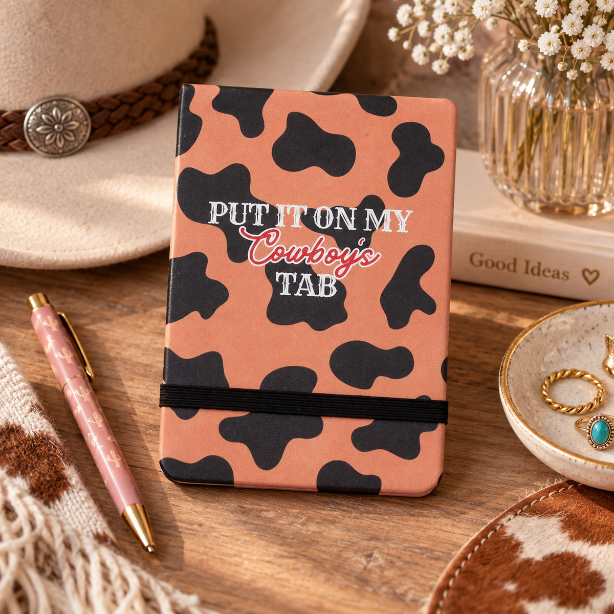 Western cowprint pocket journal styled on a rustic desk with pink pen, cowboy hat, and jewelry dish