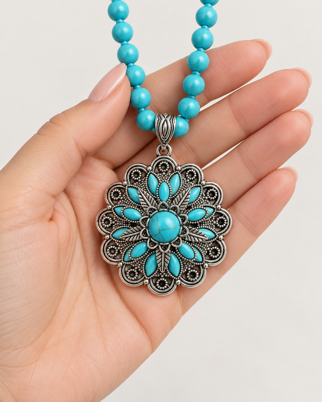 A turquoise and silver floral pendant necklace held in a hand. The necklace features a round turquoise center surrounded by a silver filigree floral pattern, with a beaded chain of turquoise beads.