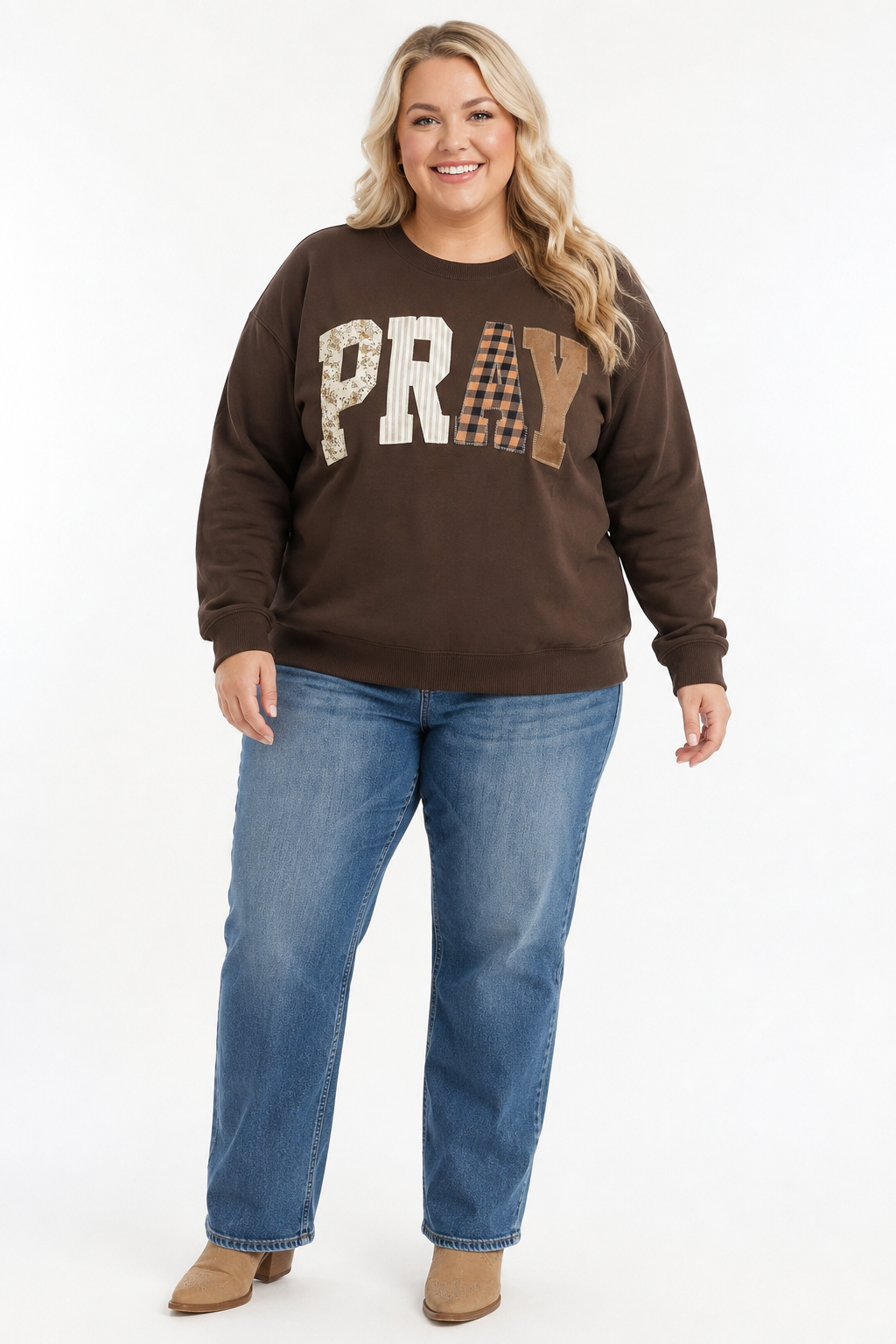 Plus-size blonde woman wearing a coffee-brown PRAY patchwork sweatshirt with jeans and tan Western boots on a white background.