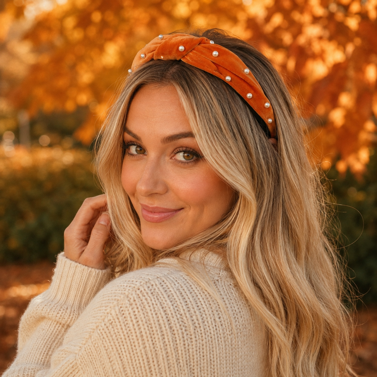 Blonde woman styling orange velvet pearl headband with cream sweater in golden autumn setting
