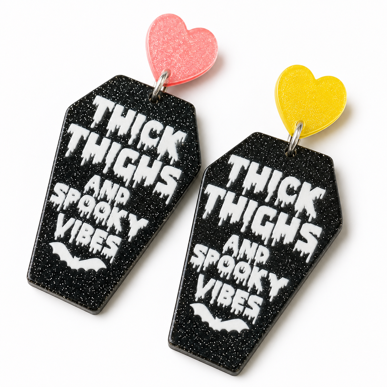 Close-up detail of Thick Thighs & Spooky Vibes Coffin Earrings showing black glitter texture, dripping white lettering, colorful heart tops, and silver connectors.