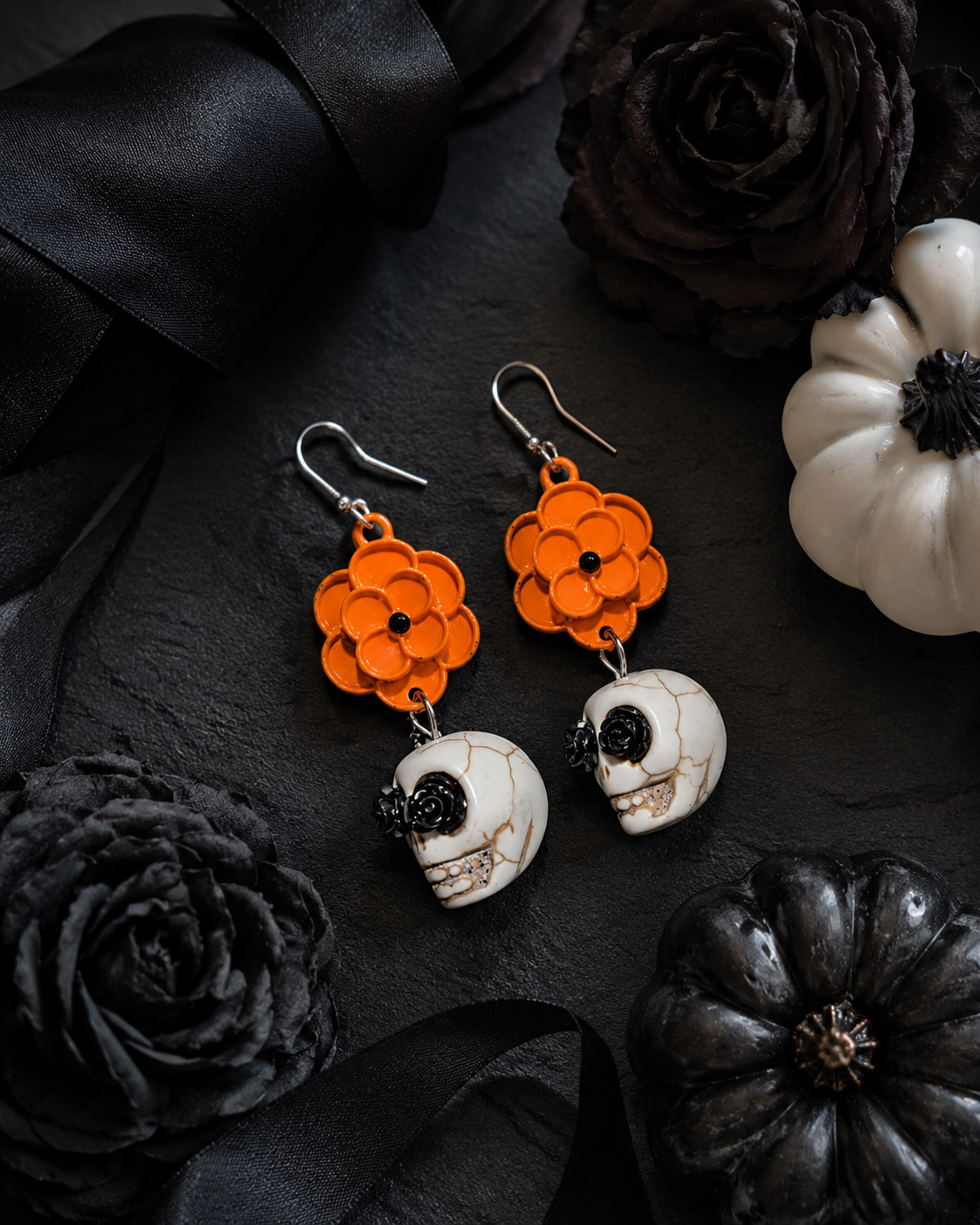Orange flower skull earrings styled with black ribbon, dark roses and Halloween pumpkins on a black background
