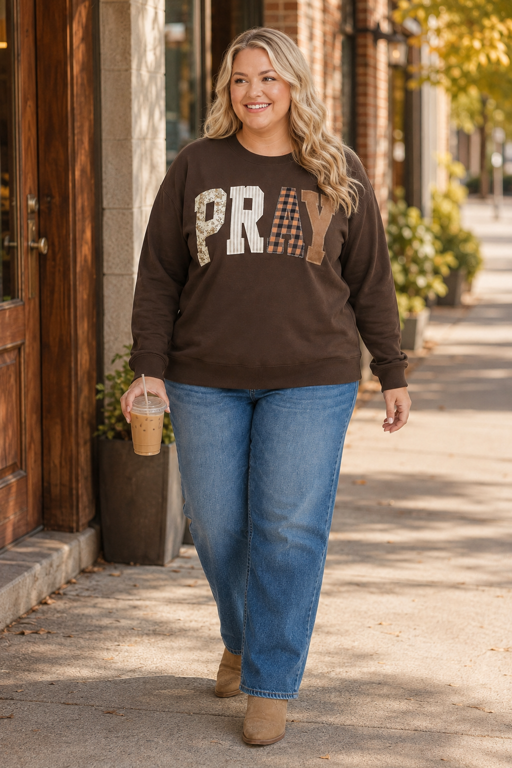 Plus-size blonde woman wearing a coffee-brown PRAY patchwork sweatshirt with jeans and Western boots while carrying iced coffee downtown.