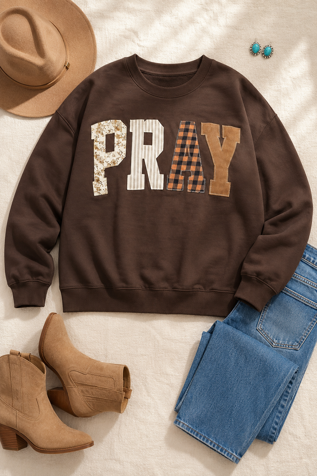 Coffee-brown PRAY patchwork sweatshirt styled with medium-wash jeans, tan Western boots, felt hat and turquoise earrings.