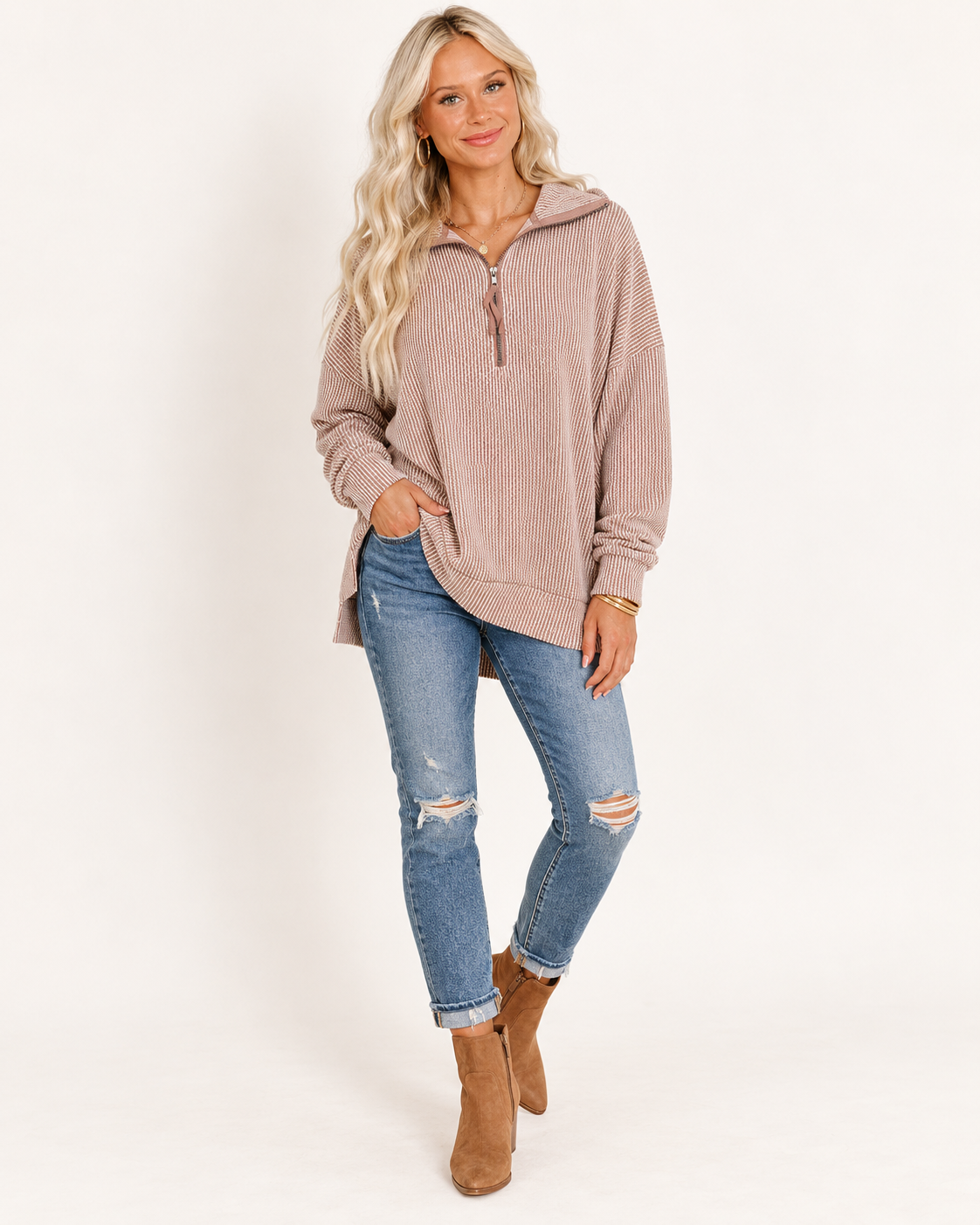 A woman modeling a light brown ribbed half-zip hoodie with a high collar, paired with distressed blue jeans and brown suede ankle boots, against a plain white studio background.