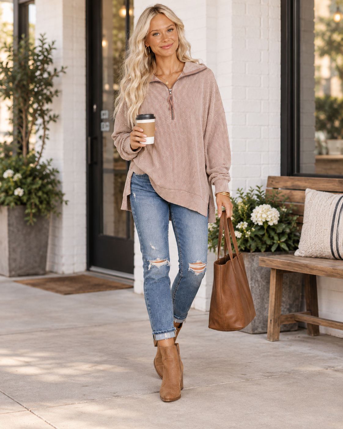 A woman stands on a sidewalk in front of a white brick building, wearing an oversized dusty rose quarter-zip hoodie, distressed blue jeans, and tan ankle boots. She holds a brown leather tote bag and a paper coffee cup.