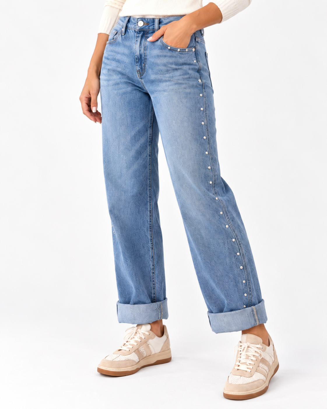A person wearing light blue denim jeans with silver stud embellishments down the outer side seams, paired with a white ribbed top and beige sneakers, photographed against a clean white studio background.