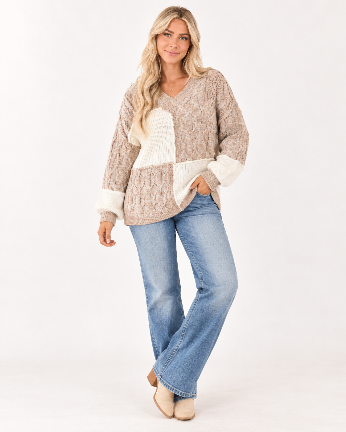 A woman modeling a beige and cream patchwork knit sweater with blue jeans and beige boots, standing against a plain white studio background.