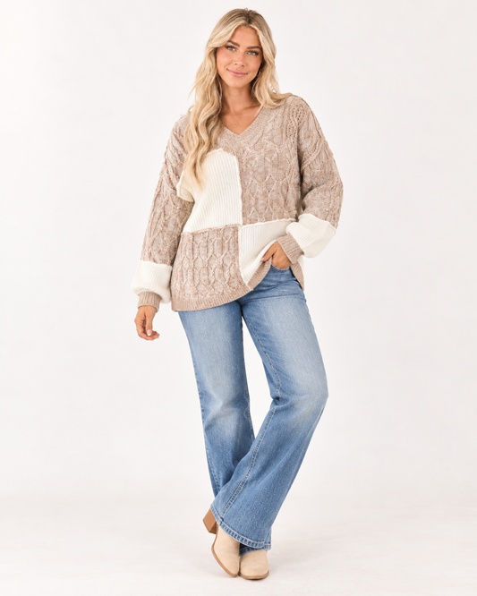 A woman modeling a beige and cream patchwork knit sweater with blue jeans and beige boots, standing against a plain white studio background.