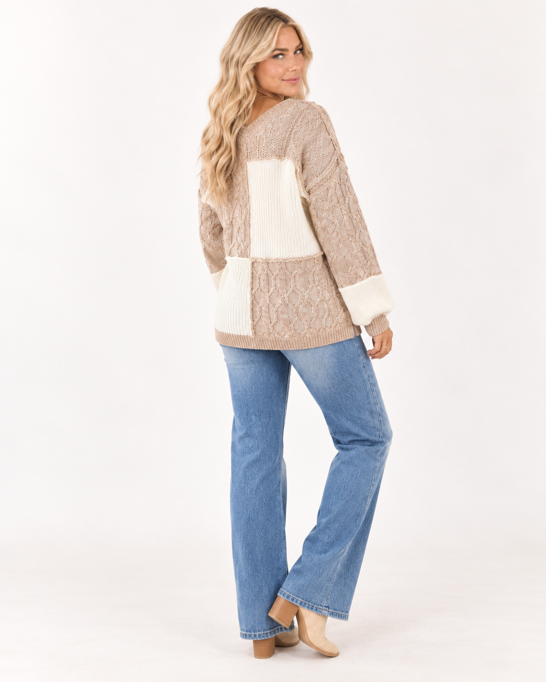 A woman modeling a chunky knit patchwork sweater in beige and cream, paired with blue jeans and beige heels, photographed against a seamless white studio background.