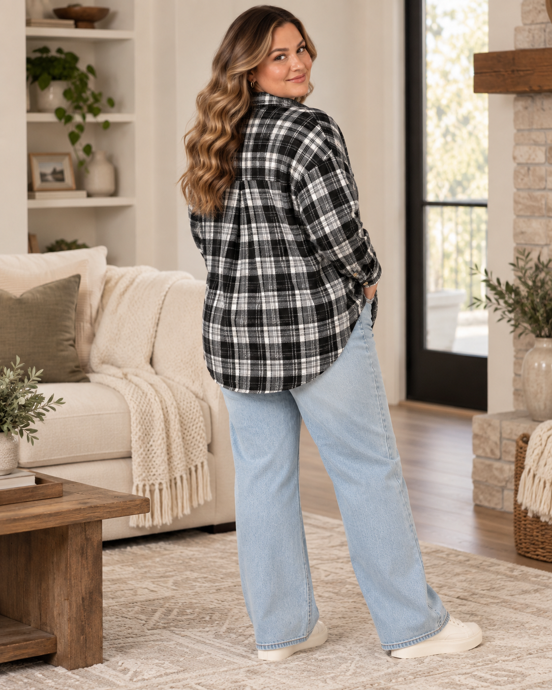 A woman stands in a living room wearing a black, white, and gray plaid button-up shirt and light blue wide-leg jeans.