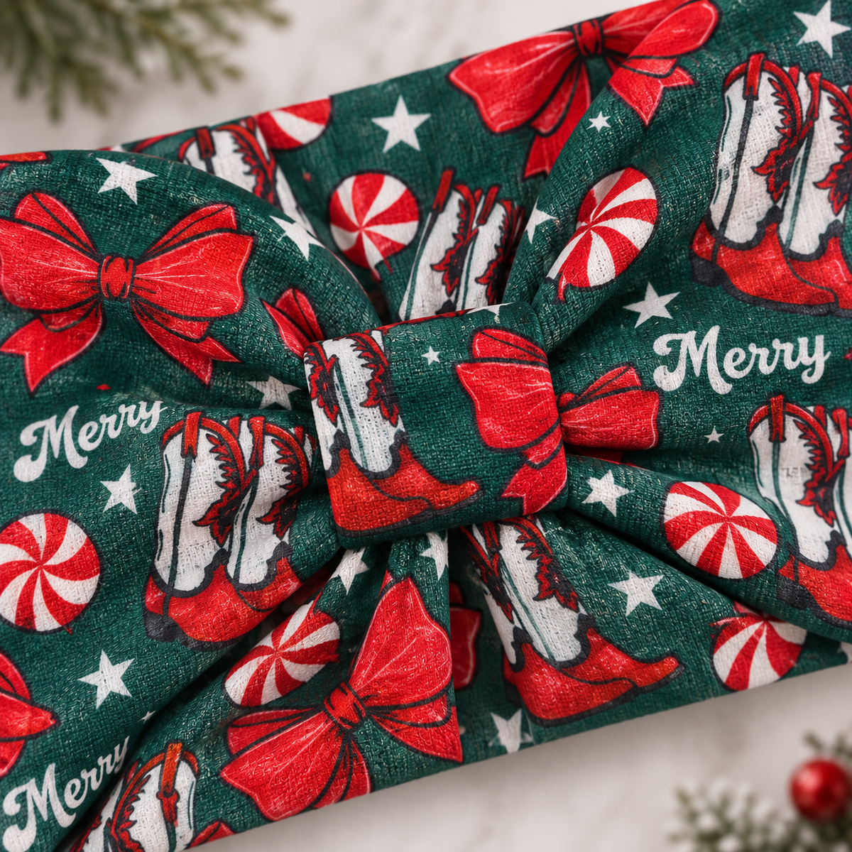 A festive Christmas-themed fabric headband with a large bow, featuring a pattern of red bows, red-and-white peppermint candies, white stars, and 'Merry' text on a dark green background.
