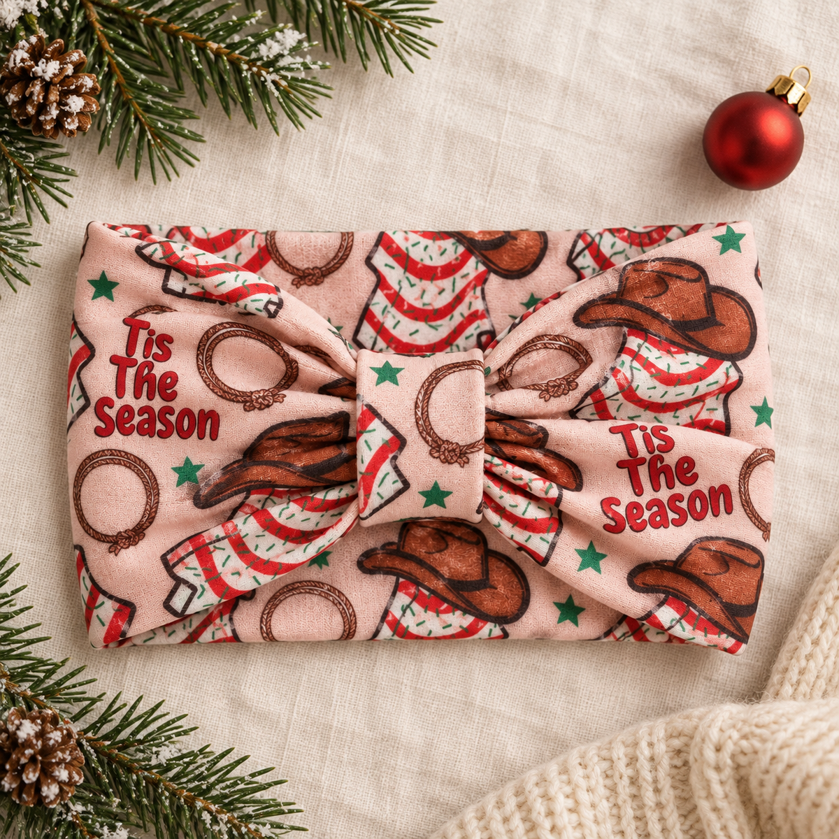 A fabric headband with a knotted center, featuring a Christmas western print with cowboy hats, lasso loops, green stars, and the text 'Tis The Season' in red. The headband is laid flat on a textured beige fabric background, styled with pine branches, a red ornament, and a knit sweater corner.