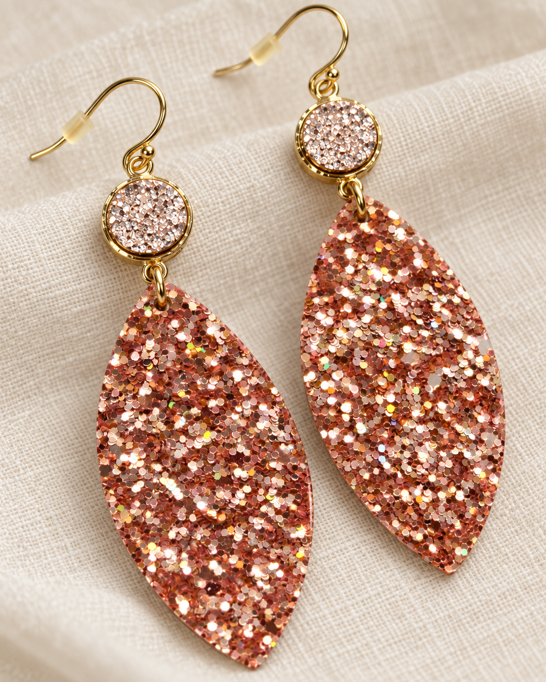 Close-up of rose-gold glitter leaf earrings styled on soft cream linen showing multicolor reflective sparkle