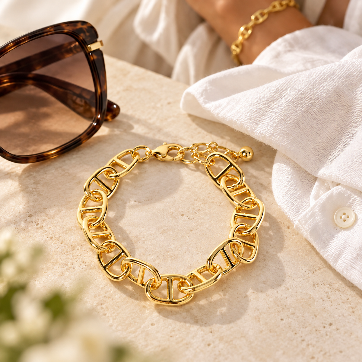 A gold chain link bracelet with a lobster clasp, styled on a beige stone surface next to tortoiseshell sunglasses and a white shirt. A person's wrist wearing a matching gold chain bracelet is visible in the upper right.