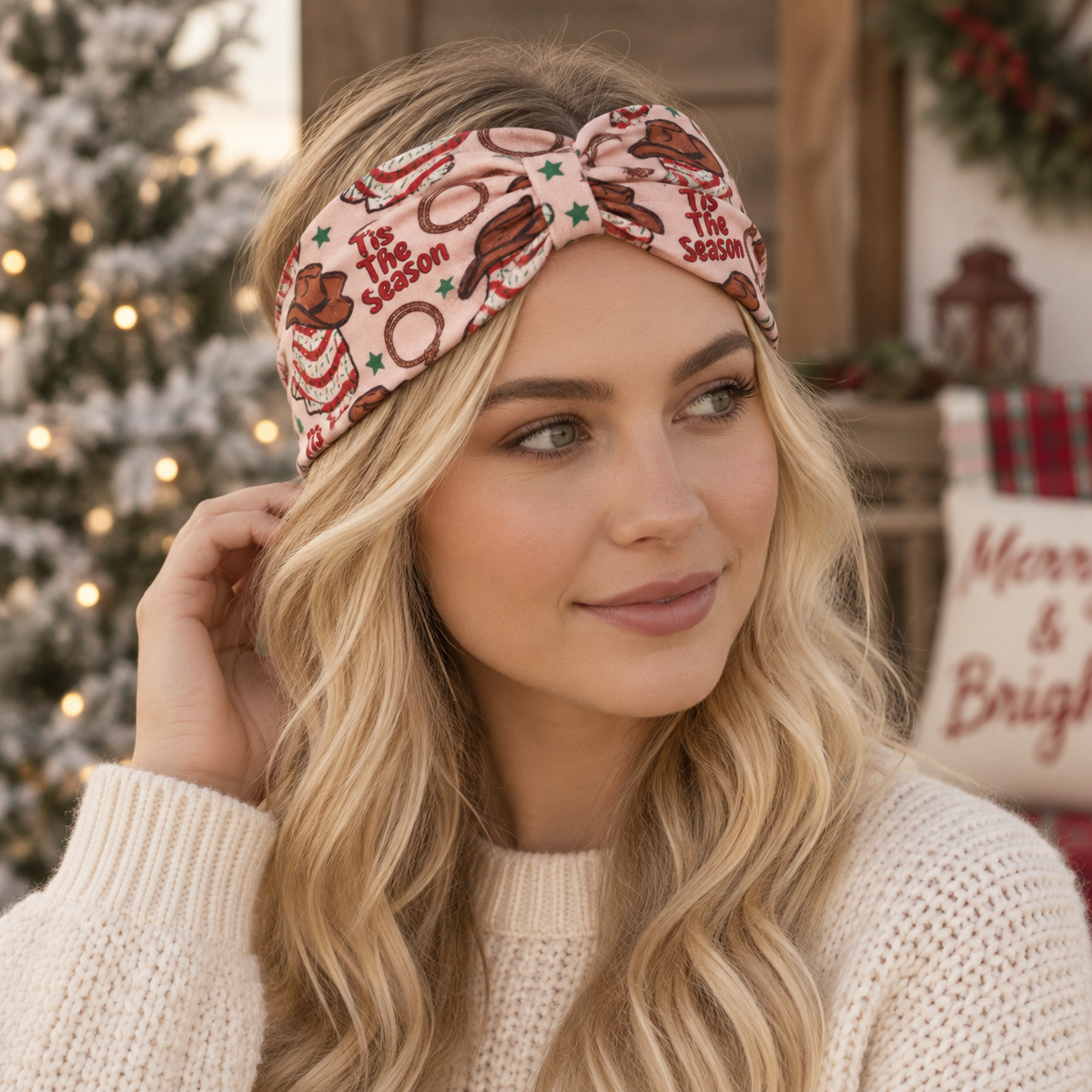A woman wearing a pink knitted holiday headband with a twisted front, decorated with gingerbread cookies, cinnamon sticks, and the words 'It's the season,' photographed in a cozy Christmas-themed setting.