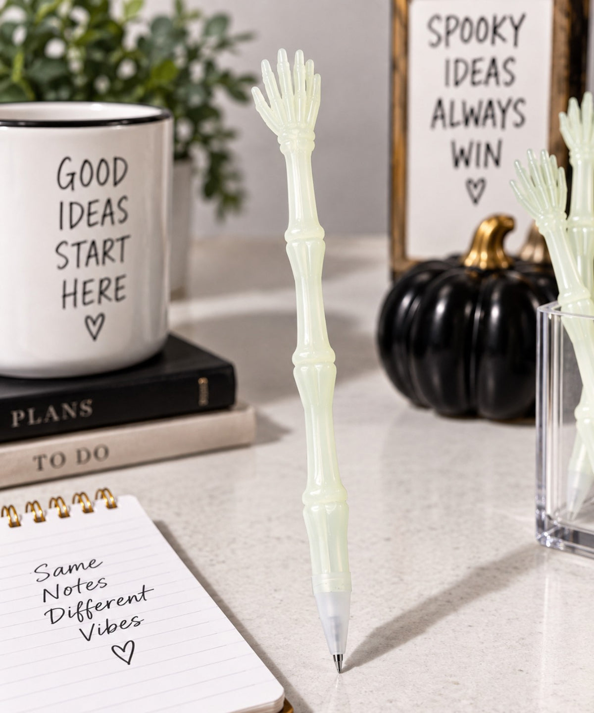 A translucent pale green pen shaped like a skeleton hand resting on a tabletop, surrounded by desk props including a mug, notebook, pumpkin, and sign.