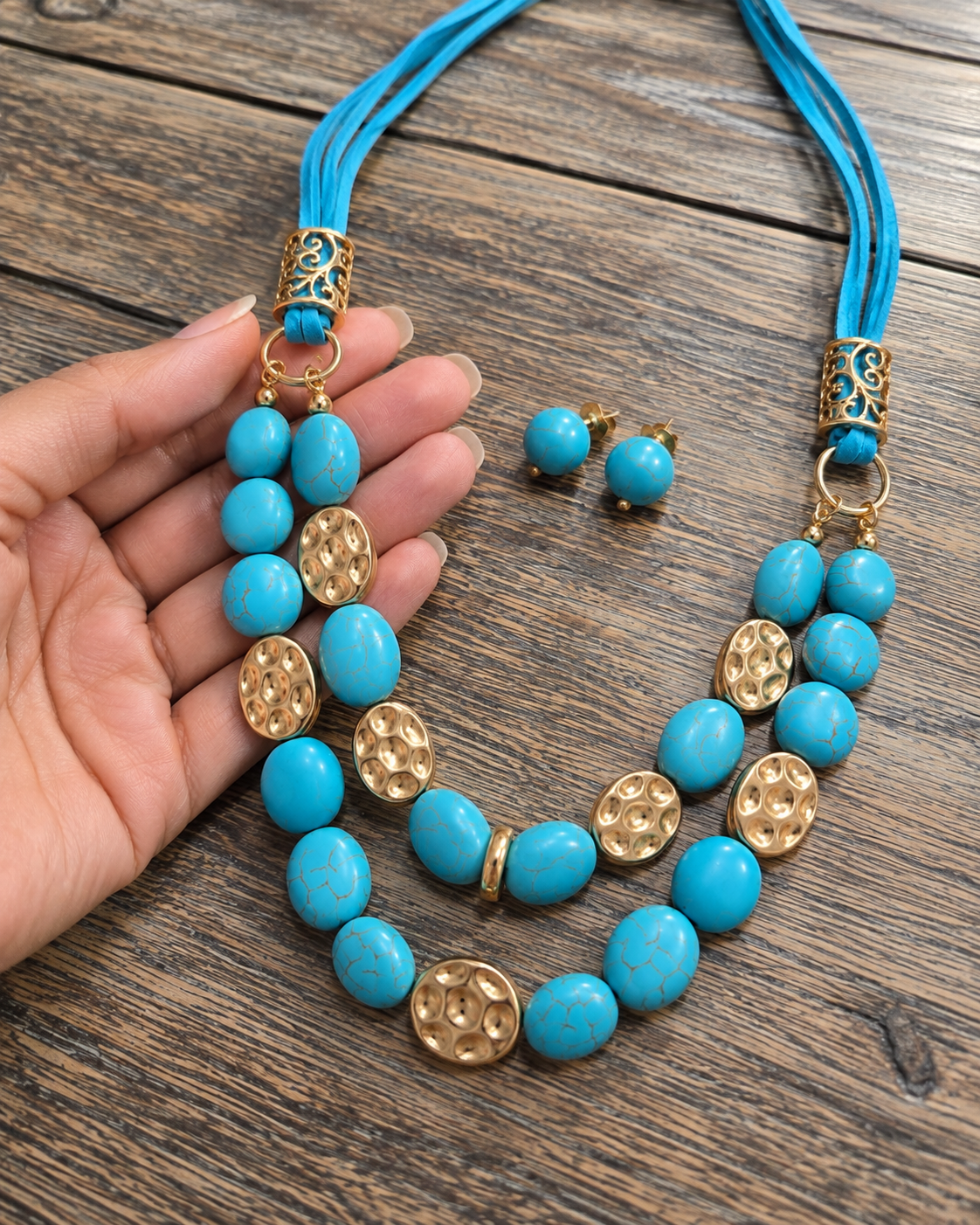 A turquoise and gold statement necklace with matching stud earrings, displayed on a rustic wooden surface with a hand holding the left side of the necklace.