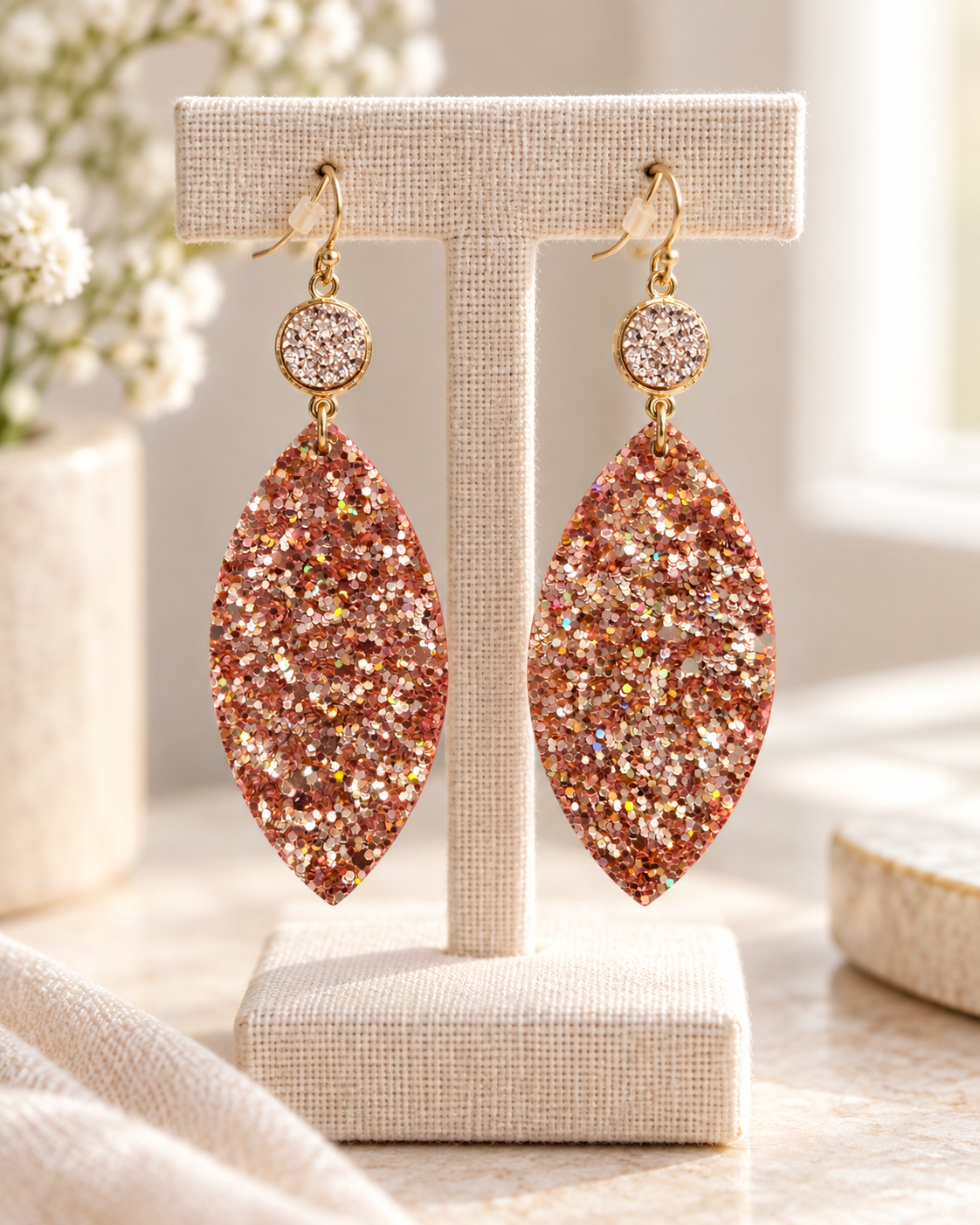 Rose-gold glitter leaf earrings displayed on a neutral linen jewelry stand in soft natural light