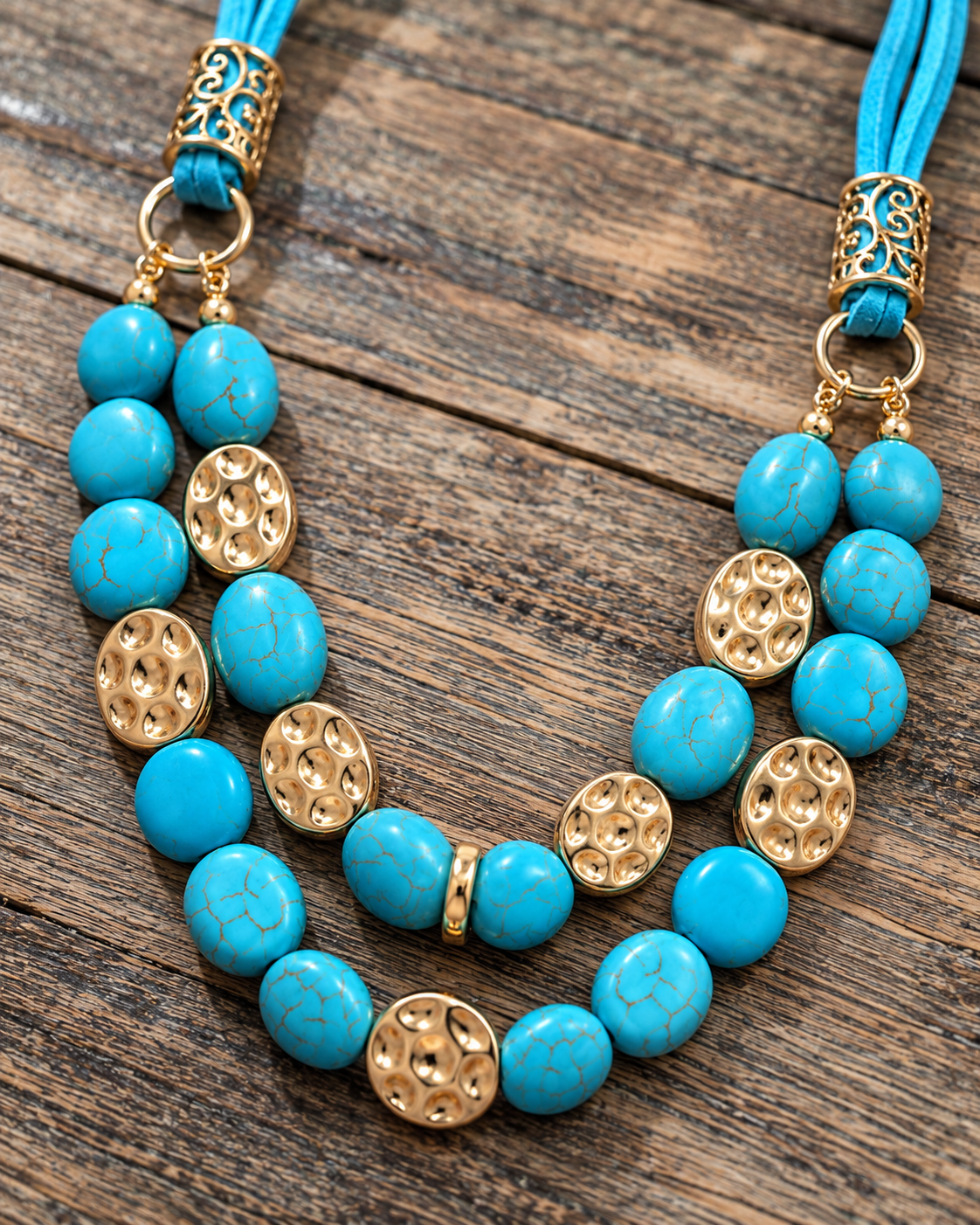 A double-strand turquoise and gold beaded necklace laid flat on a rustic wooden surface. The necklace features round turquoise beads, textured gold disc beads, and gold filigree cap details with turquoise cords extending to the top of the frame.