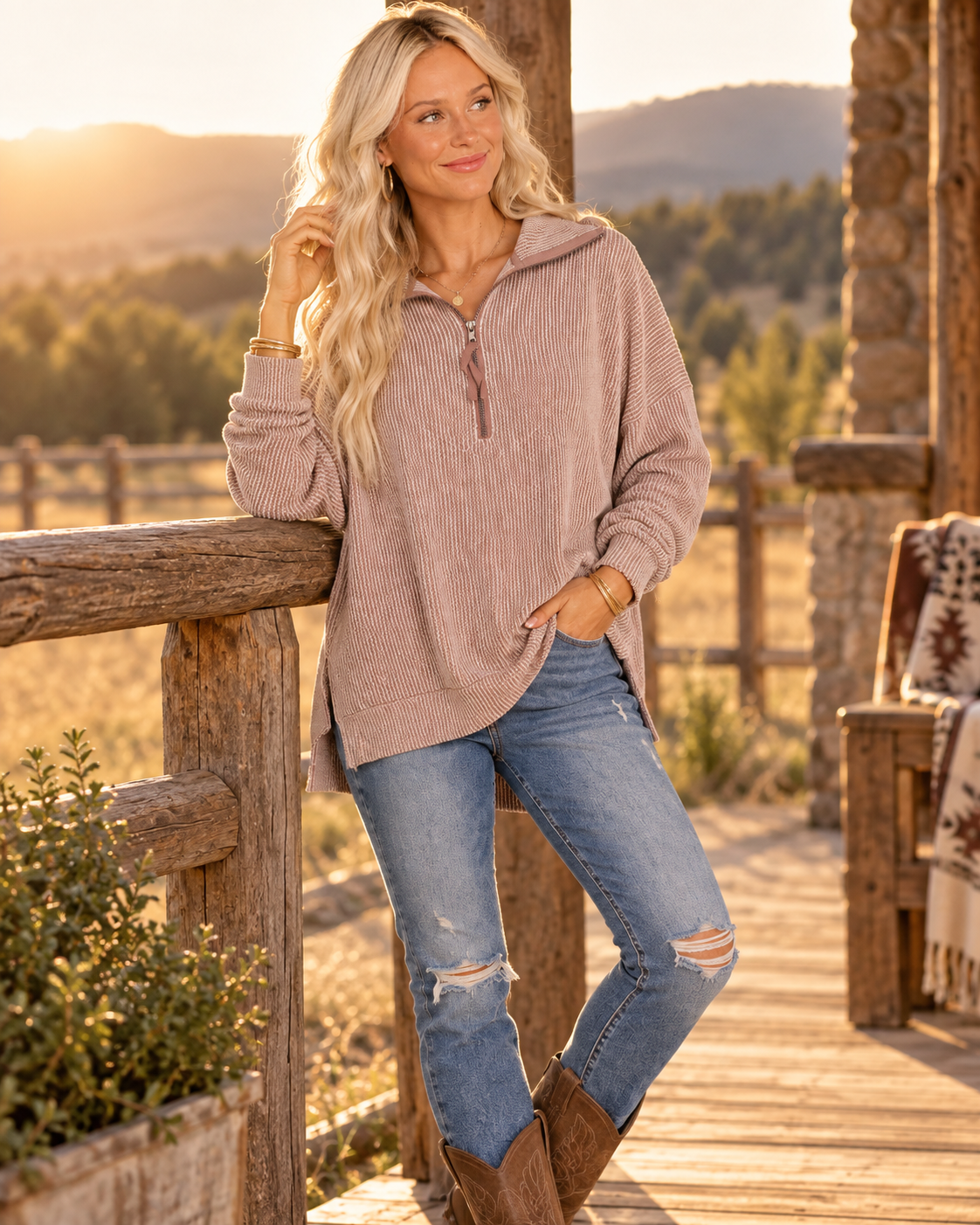 A woman stands on a rustic wooden porch wearing a ribbed, dusty mauve-pink quarter-zip pullover sweater, distressed blue jeans, and brown cowboy boots, with a warm outdoor mountain landscape in the background.