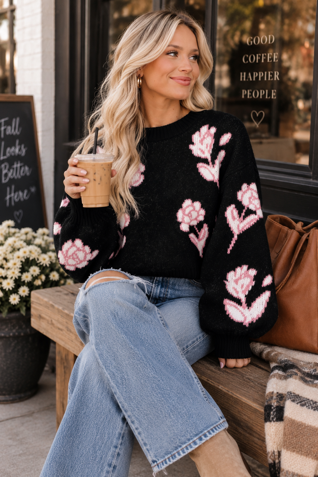 A woman sits on a wooden bench outside a coffee shop, wearing a black knit sweater with pink floral patterns and light blue jeans, holding a coffee cup.