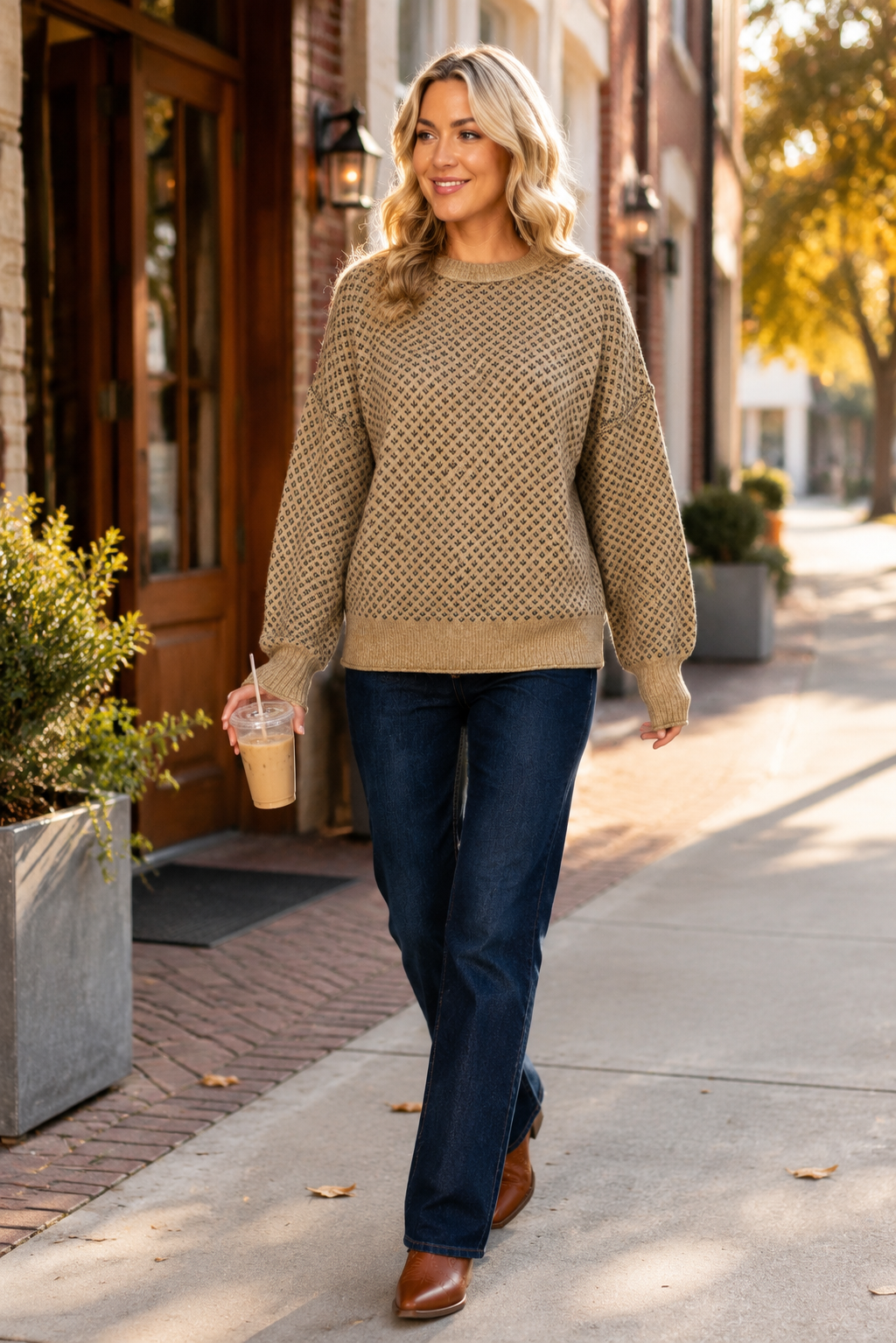 Woman wearing a khaki geometric sweater with dark jeans and boots while carrying iced coffee through a historic downtown.