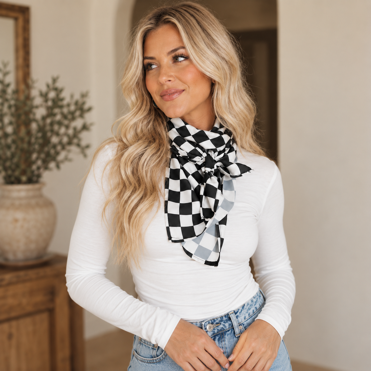 A woman with blonde hair wearing a black and white checkered scarf tied around her neck, a white long-sleeve top, and blue jeans, standing in a warmly lit indoor setting.