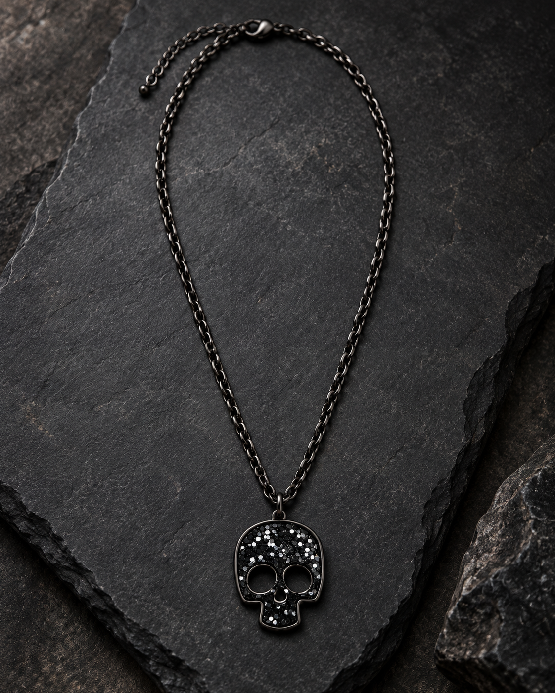 A gothic-style skull pendant necklace with a dark oxidized metal finish, featuring small clear rhinestones on the forehead, displayed on a dark slate background.