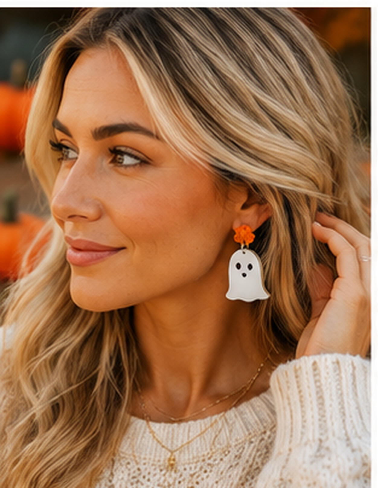 A woman wearing a white ghost-shaped dangle earring with a small orange flower accent, shown in a close-up portrait against a softly blurred autumn background.