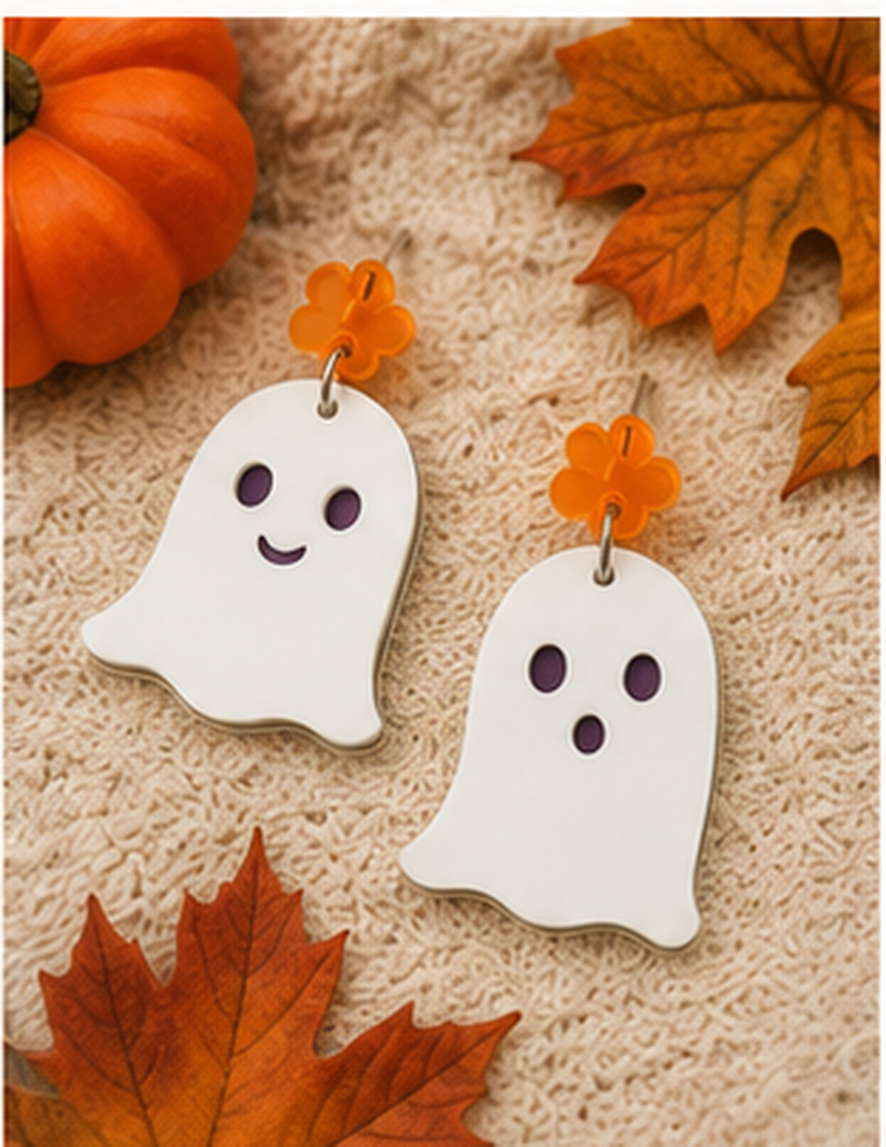 A pair of white ghost-shaped dangle earrings with purple eyes and smiles, hanging from bright orange flower charms, displayed on a beige textured fabric with autumn leaves and a small pumpkin.