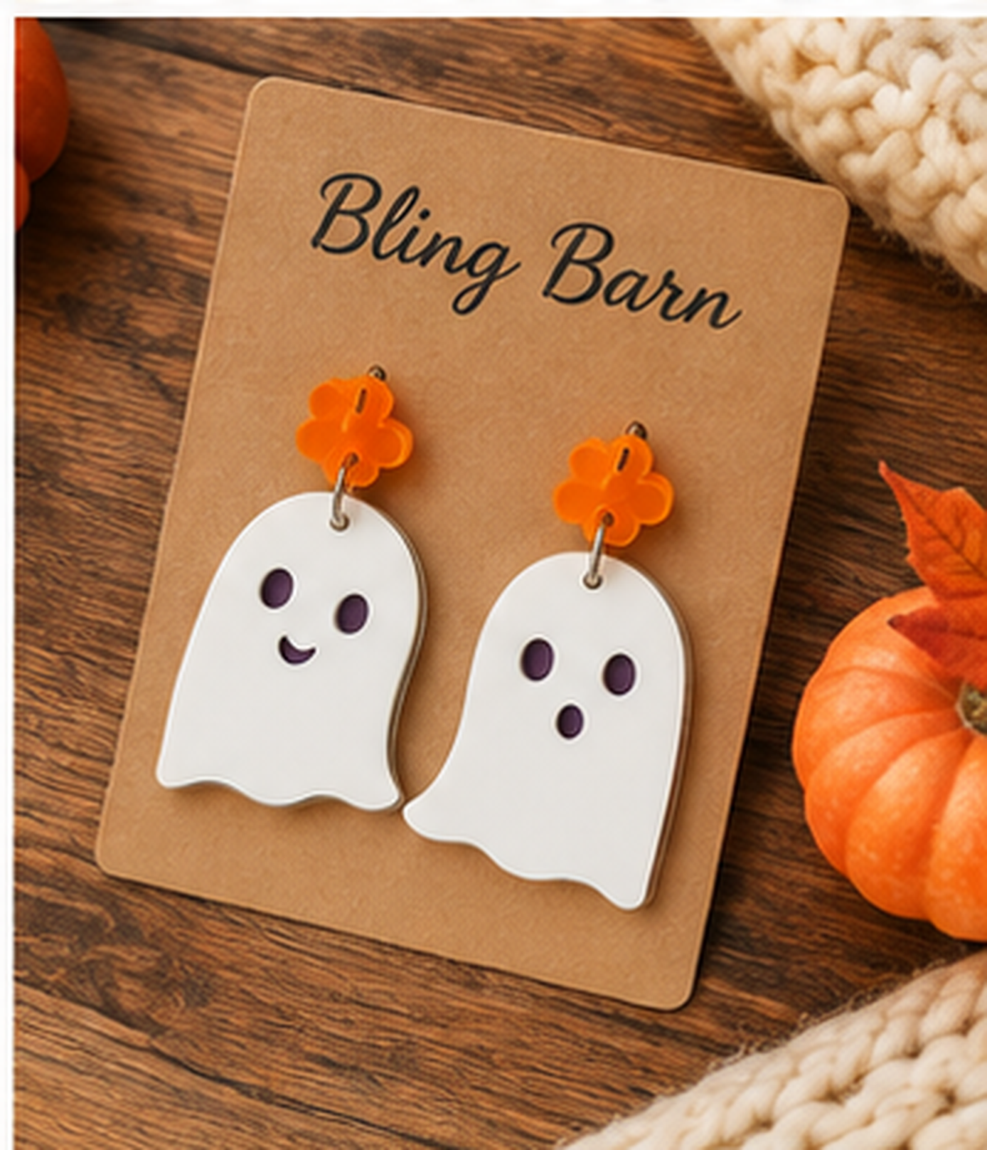 A pair of white ghost-shaped dangle earrings with orange flower studs, displayed on a brown card labeled 'Bling Barn' on a wooden surface with autumn props.