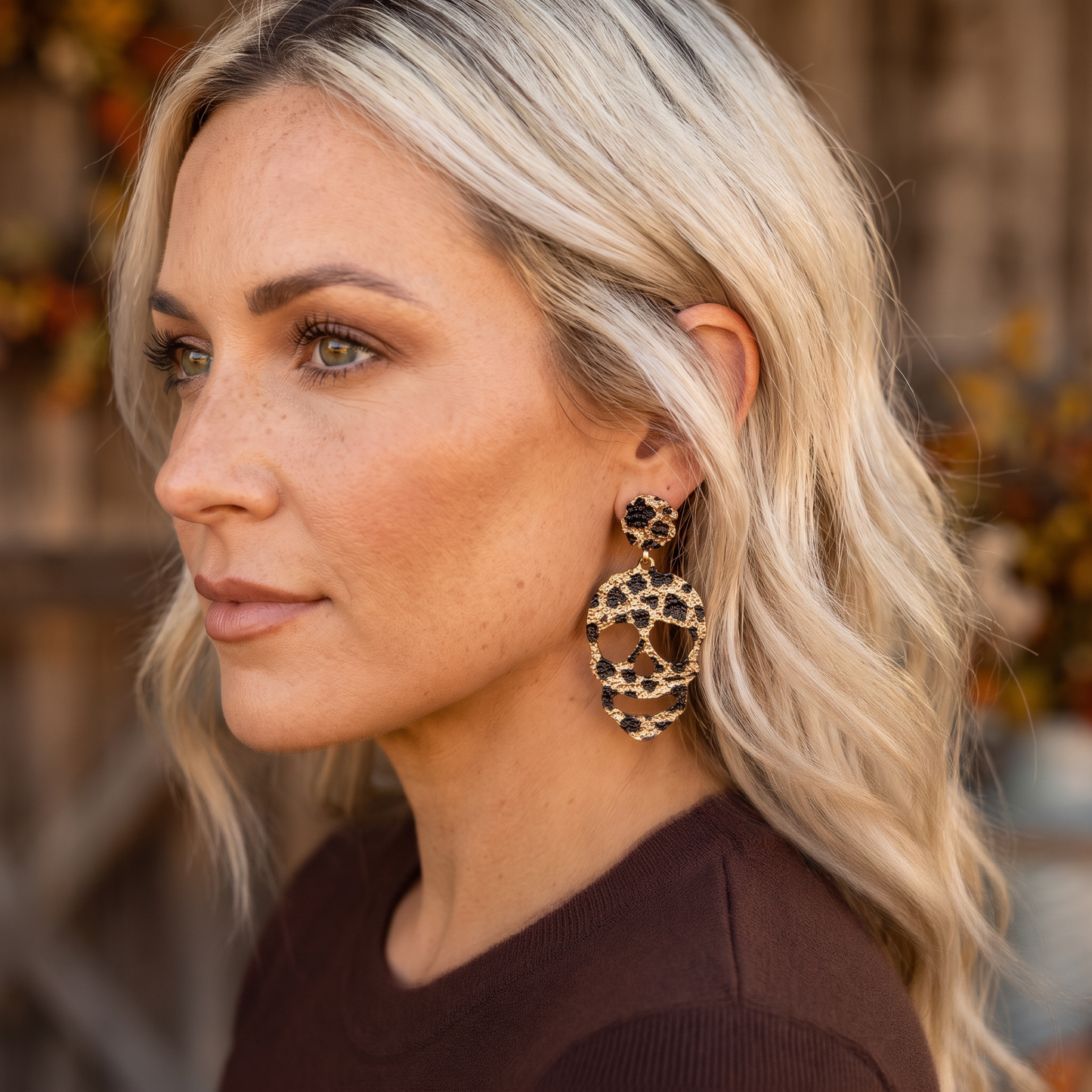 A close-up side profile of a woman with blonde hair wearing a large, dangling statement earring with a gold-tone circular frame and black leopard-print spots, set against a blurred outdoor background.