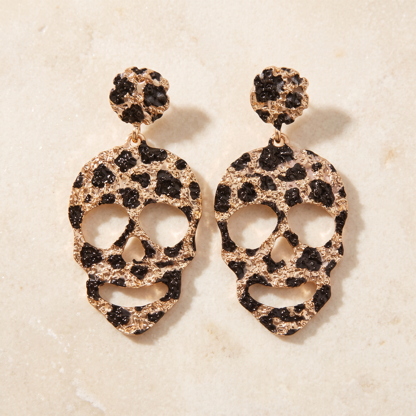 A pair of dangling skull-shaped earrings with a textured leopard print pattern in black and rose gold, displayed on a light beige stone surface.