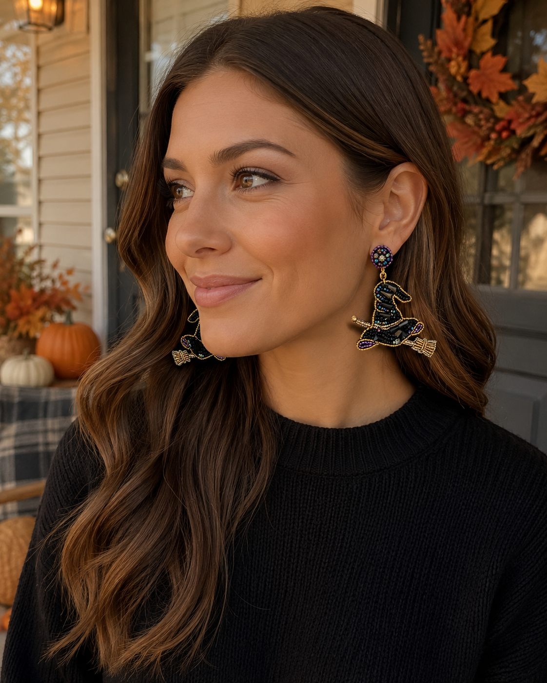 Woman wearing beaded witch hat earrings with a black sweater in a warm autumn porch setting