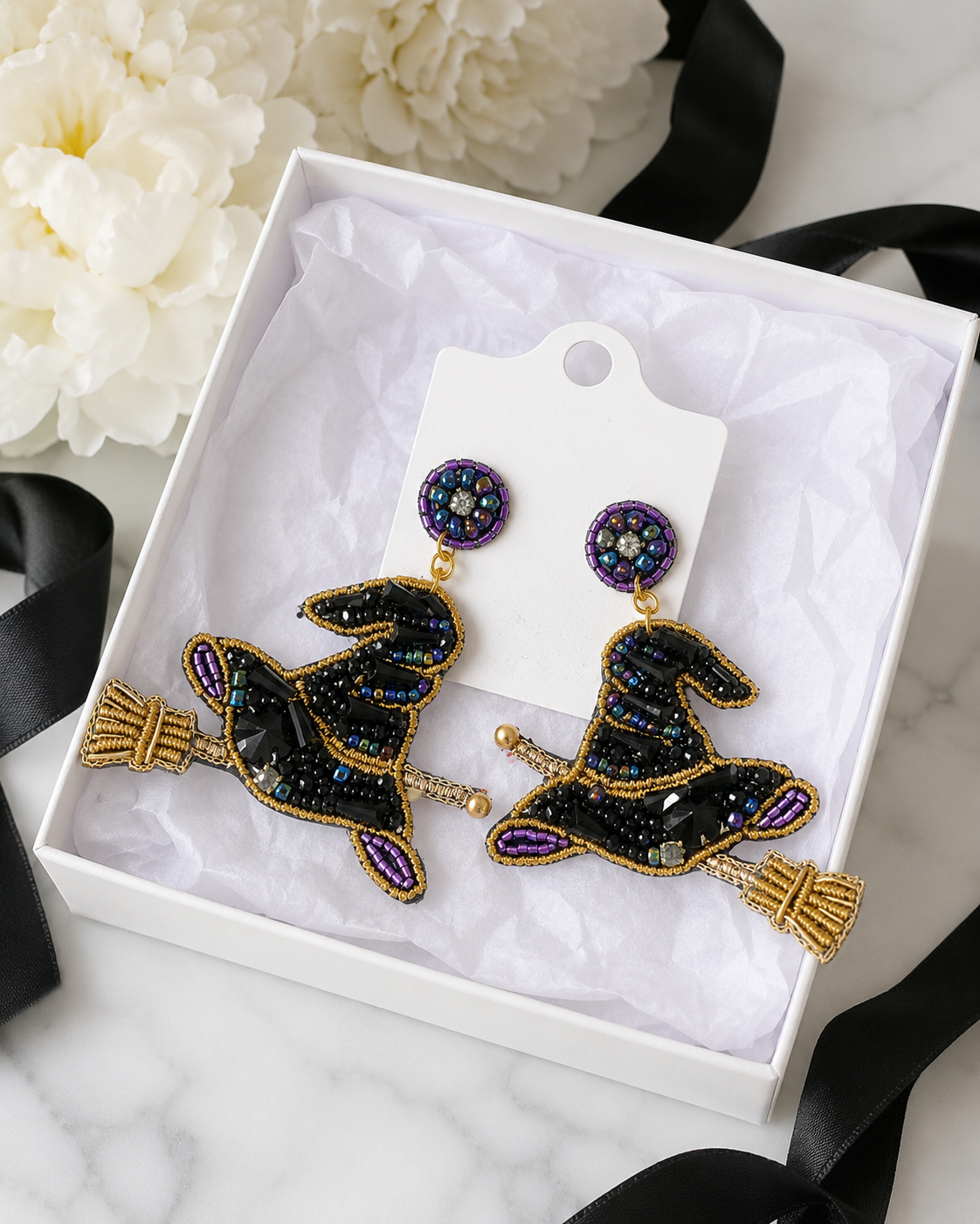 Beaded witch hat and broom earrings displayed on a white jewelry card inside a gift box with black ribbon