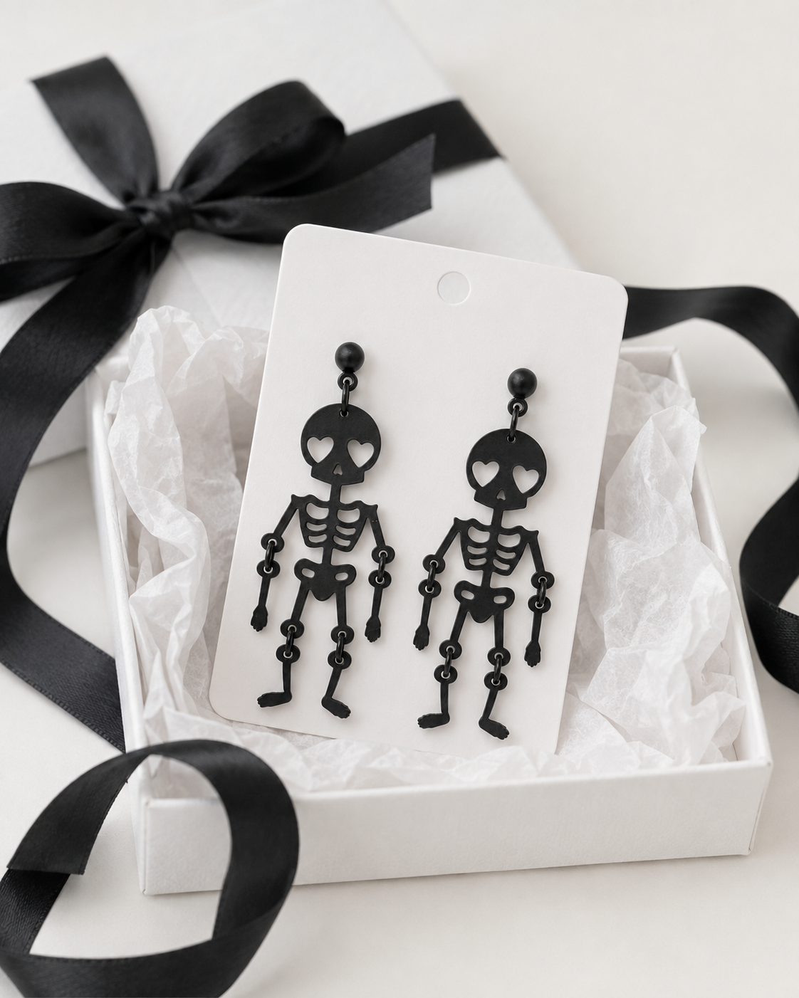 Love You to Death skeleton earrings displayed on a white jewelry card with gift box, tissue and black ribbon