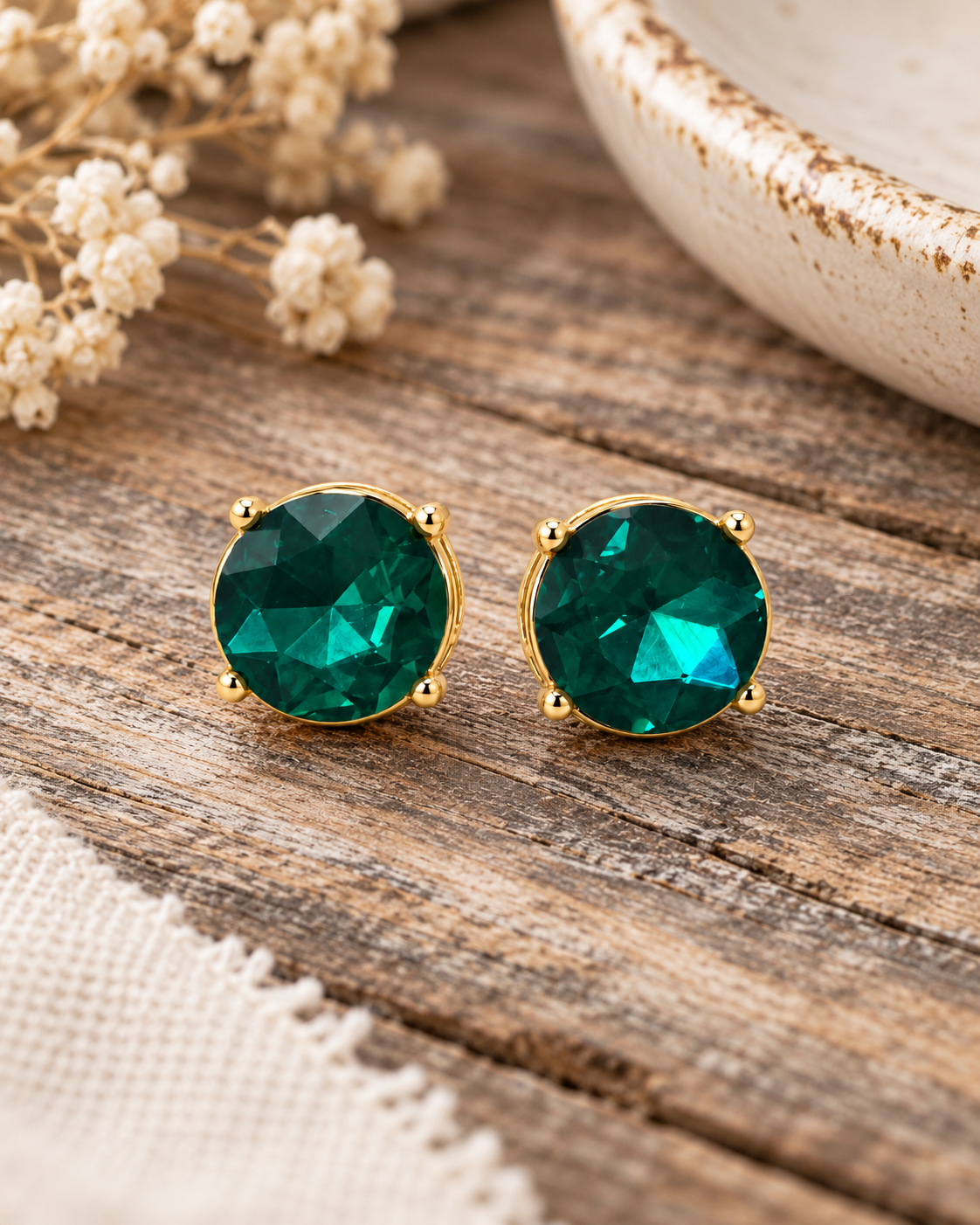 A pair of round green faceted stud earrings with gold settings, displayed on a rustic wooden surface with dried flowers and a ceramic bowl in the background.