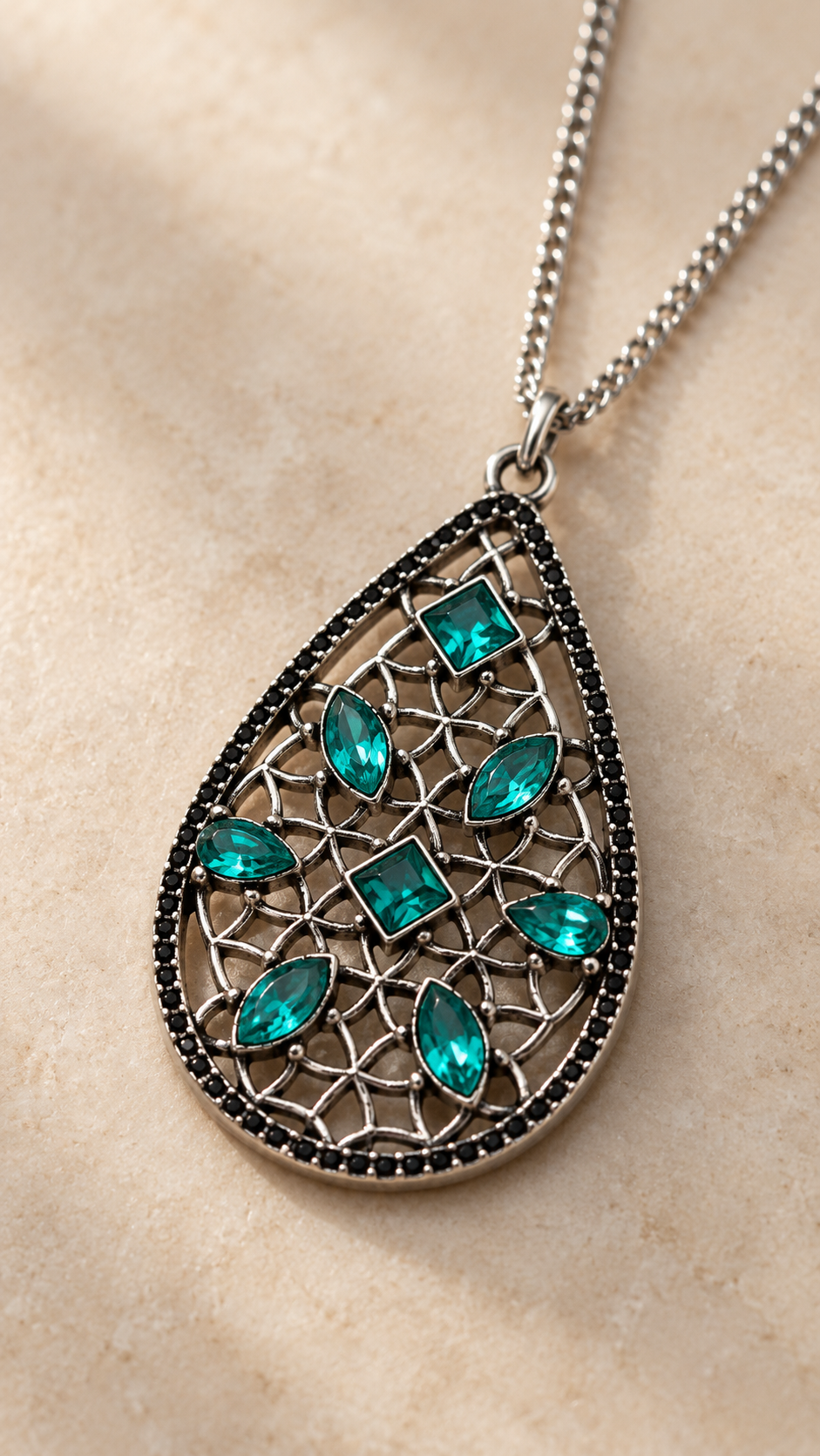 A teardrop-shaped silver pendant necklace with a filigree design, featuring seven sparkling teal green gemstones, displayed on a textured beige surface.