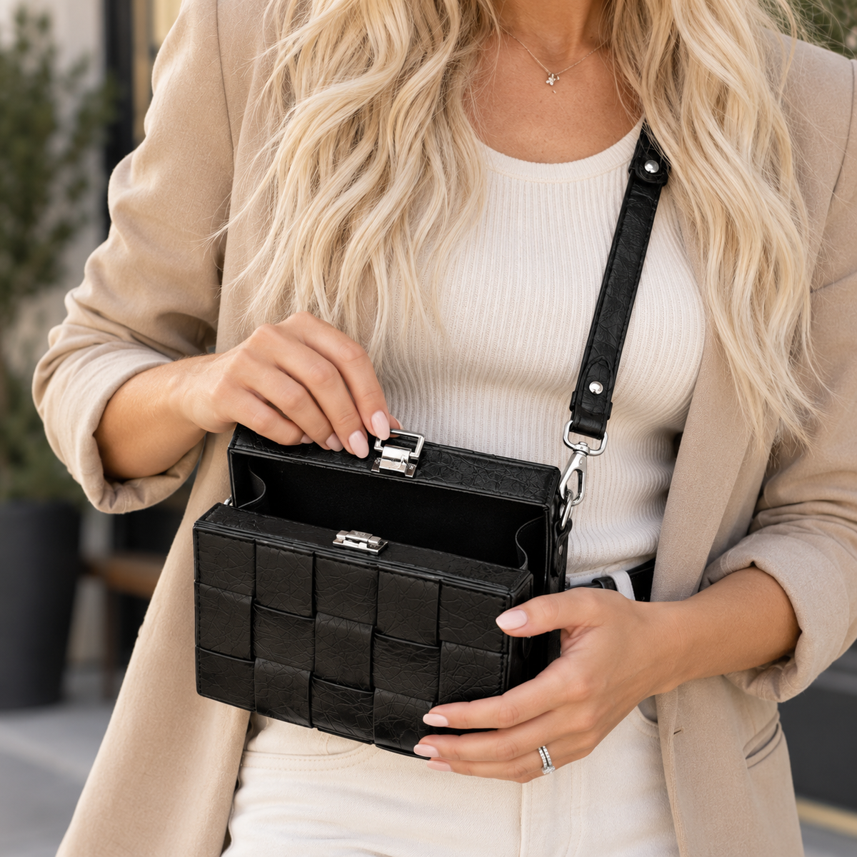 A woman wearing a beige blazer and cream ribbed top holds a black woven leather clutch bag with a silver clasp and black shoulder strap.