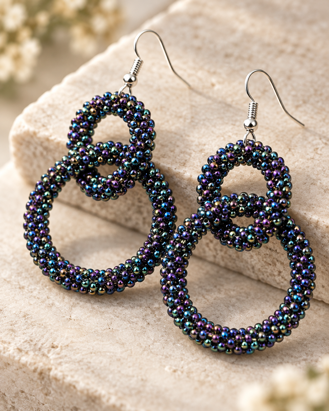 A pair of large, double-loop dangle earrings made from iridescent seed beads that shift between blue, green, and purple, hanging from silver fishhook ear wires. They are displayed on a textured beige stone block with soft, out-of-focus flowers in the background.