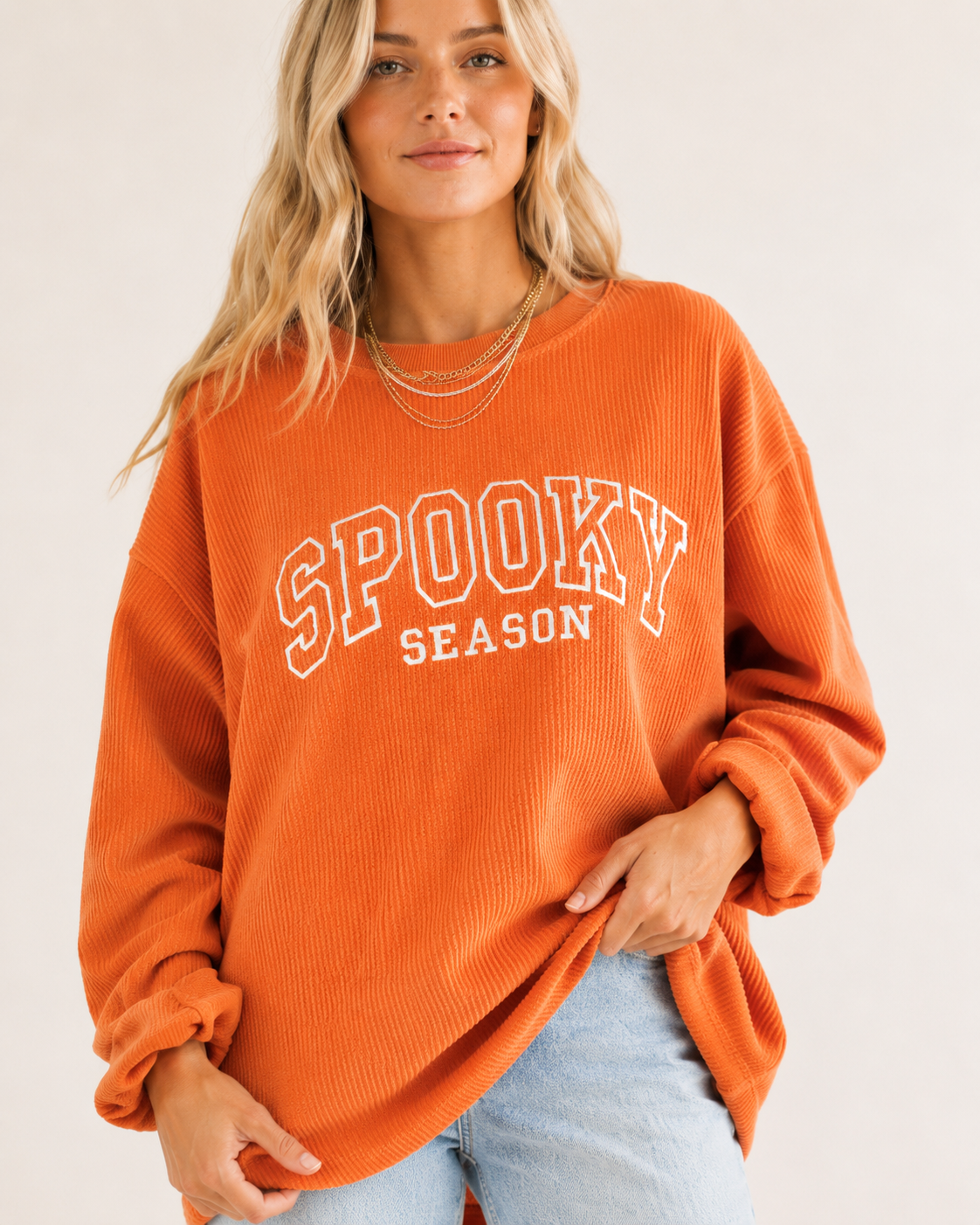 A woman wearing an orange ribbed crewneck sweatshirt with 'SPOOKY SEASON' printed in white arched letters on the front, paired with light blue jeans against a plain off-white background.